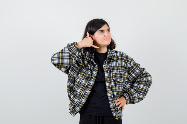 Portrait of teen girl showing phone gesture, keeping hand on waist in casual shirt and looking blissful front view