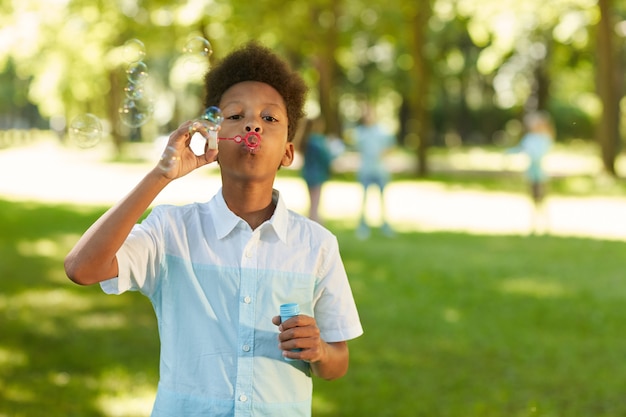 Portrait of teenage African-American boy blowing bubbles while standing in green park outdoors