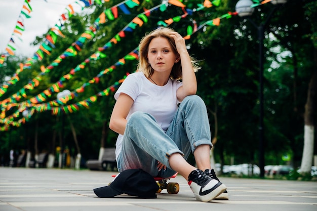 Portrait of a teenage girl in casual clothes in the park Summer skateboarding active lifestyle A student or a schoolboy is sitting on a skateboard during the summer holidays Sports recreation