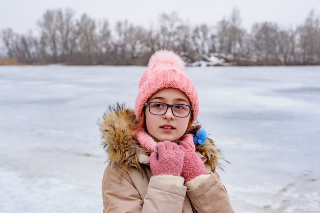 Portrait of teenage girl in pink hat and coat standing in winter landscape. cute girl with glasses