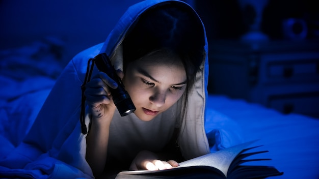 Portrait of teenage girl reading book in bed with flashlight.