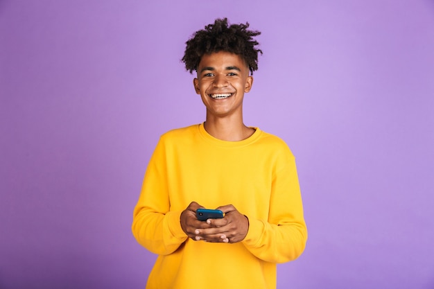 Portrait of teenager boy with afro hairdo smiling and holding smartphone, wearing bluetooth earphone, isolated over violet background