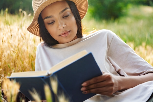 Portrait of thinking woman wearing lip piercing and straw hat reading book while lying on grass in green park