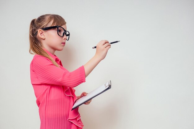 Portrait of thoughtful little smiling girl in a pink blouse and glasses holding notebook and pencil up with tablet copyspace. genius child with the idea on white background in studio copy space