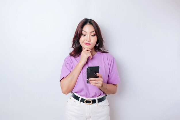 Portrait of a thoughtful young Asian woman wearing lilac purple tshirt looking aside while holding smartphone