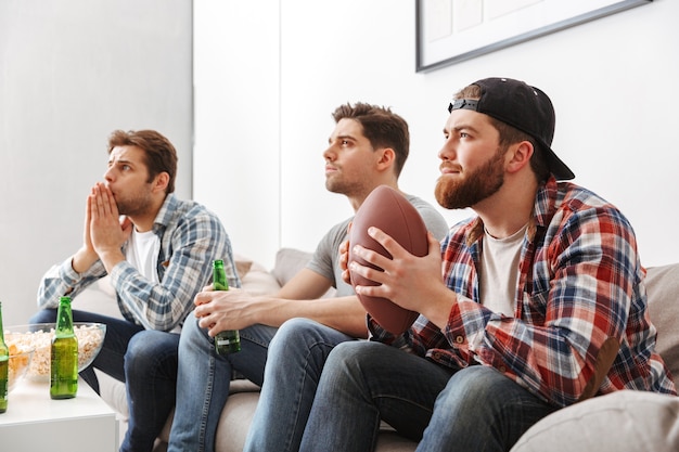 Portrait of three concentrated young men watching american football while sitting at home with beer and snacks indoors