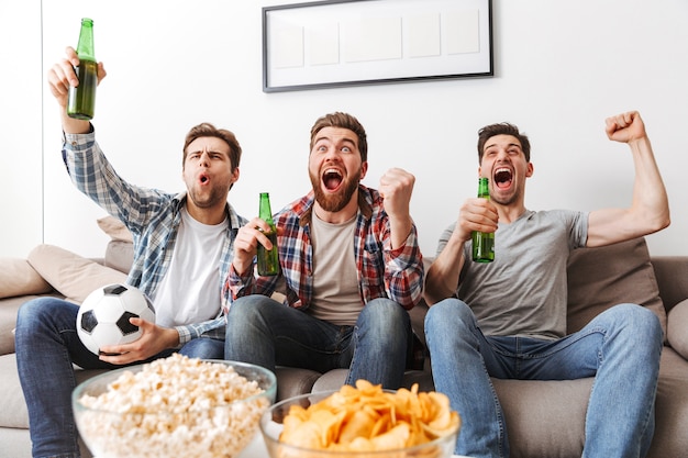 Portrait of three happy young men watching football while sitting at home, drinking beer and eating snacks