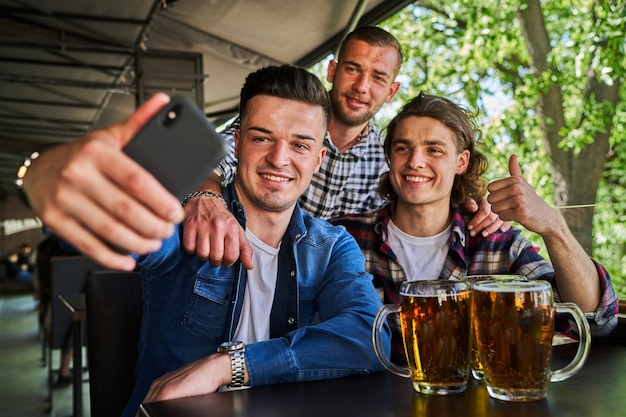 Portrait of three male friends drinking beer and make selfie in pub.