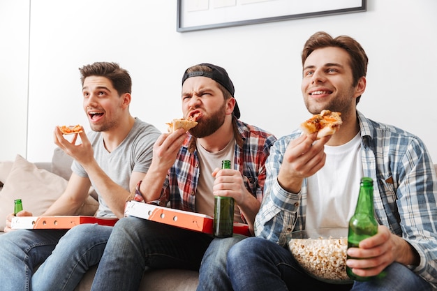 Portrait of three young bachelors eating pizza with pleasure, while watching football match at home tv