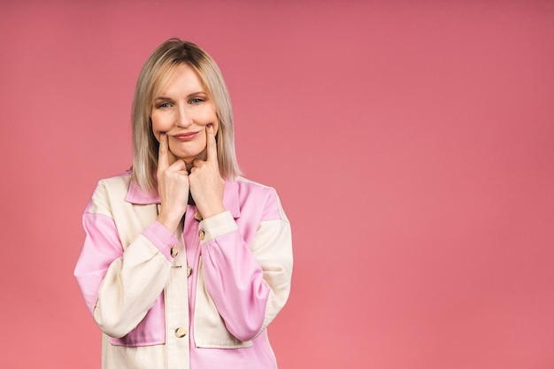 Portrait of a tired middle-aged female blonde woman has a terrible headache, feels pain and frustration, has an unhappy expression, isolated over pink background.