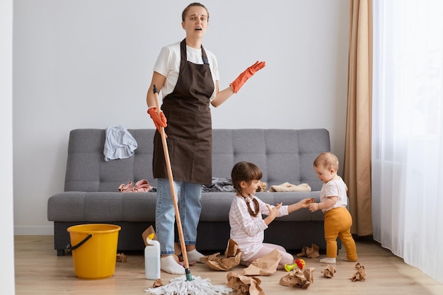 Portrait of tired mother wearing brown apron cleaning at home woman with mop cleaning with kids sitting on floor at the sofa exhausted housewife of tidying up house