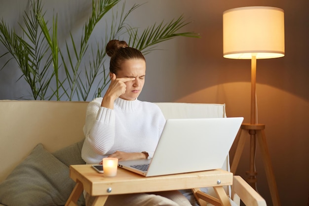 Portrait of tired sad sleepy woman wearing white sweater sitting on sofa and working on notebook feels pain in her eyes rubbing her eye feeling fatigue