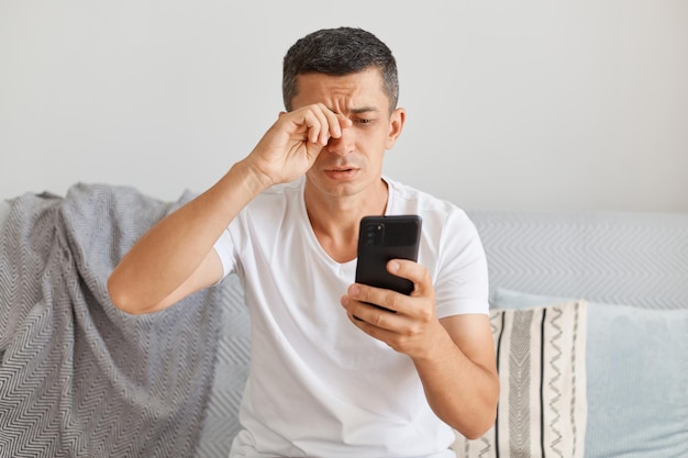 Portrait of tired upset man wearing casual style white t shirt sitting on cough using smart phone looks exhausted rubbing eye looking at gadget screen with sorrow