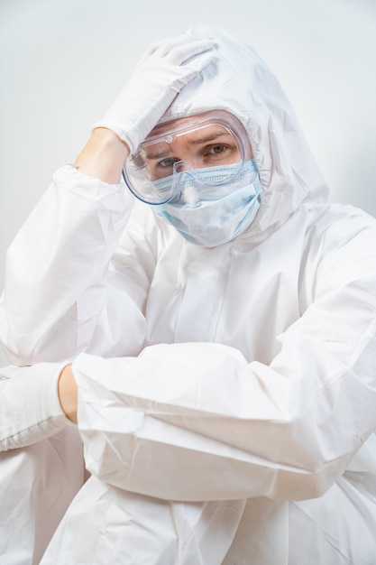 Portrait of a tired woman doctor in a protective suit, glasses, mask and glove against the white wall.