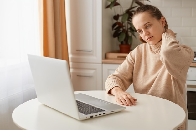 Portrait of tired woman with ponytail hairstyle wearing casual style beige sweater sitting at table in kitchen and working online on computer, feels pain in neck after long hour work, looks exhausted.