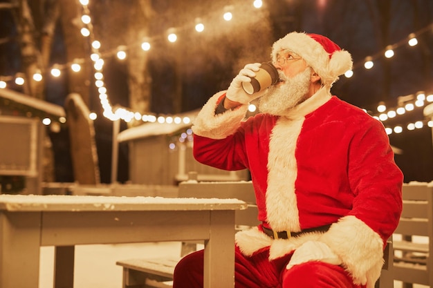 Portrait of traditional santa claus drinking hot cocoa in park with fairy lights in background copy