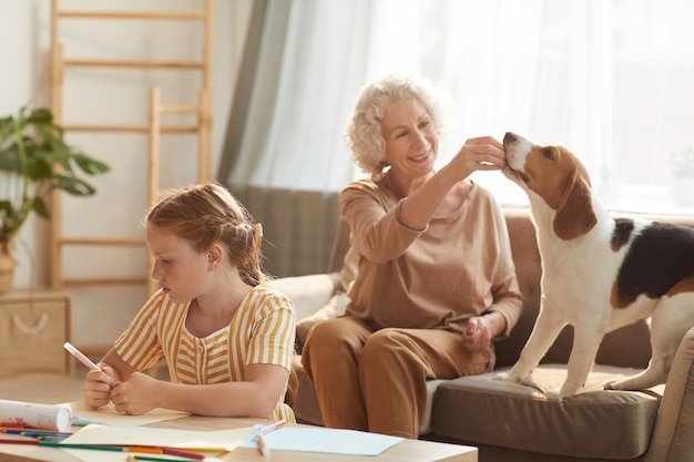 portrait of tranquil family scene with senior woman playing with dog and cute red haired girl drawing pictures beside her in cozy home interior