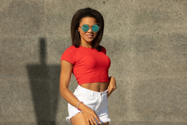 Portrait of trendy young black woman in white shorts and red top walking outdoors.