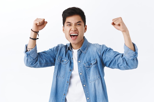 Portrait of triumphing successful asian man fist pump rejoicing yelling yes hooray at camera winning celebrating victory being excited of team scored goal rejoicing over white background