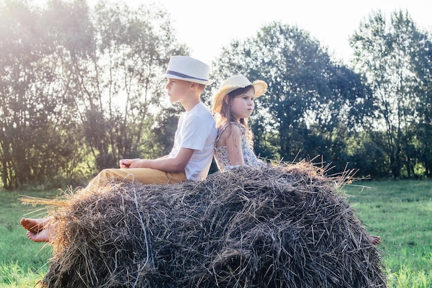 Portrait of two barefoot children boy and girl sitting their backs to each other on haystack in field Kids wearing hats