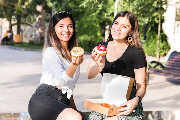 Portrait of two beautiful girls friends enjoying delicious donuts with caramel and raspberry in city park. junk but tasty food for positive mood.