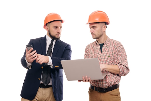 Portrait of two builders in protective orange helmets standing on white isolated background and looking at laptop display. Discuss construction project