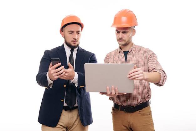 Portrait of two builders in protective orange helmets standing on white isolated background and looking at laptop display. Discuss construction project