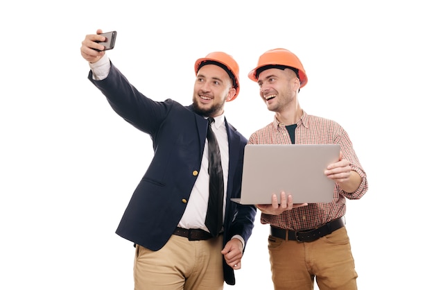 Portrait of two builders in protective orange helmets standing on white isolated background and looking at laptop display. Discuss construction project