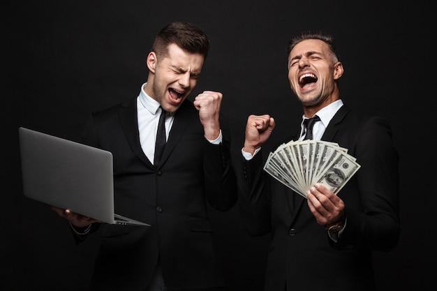 Portrait of two businessmen dressed in formal suit celebrating while holding laptop and money banknotes isolated over black wall