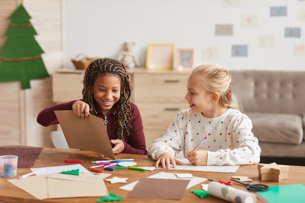 Portrait of two cheerful girls enjoying crafting and painting toether while siting at desk in decorated playroom