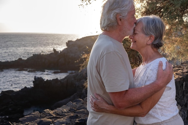 Portrait of two happy and romantic seniors or pensioners embraced at the sea at sunset light old people outdoors enjoying vacations together