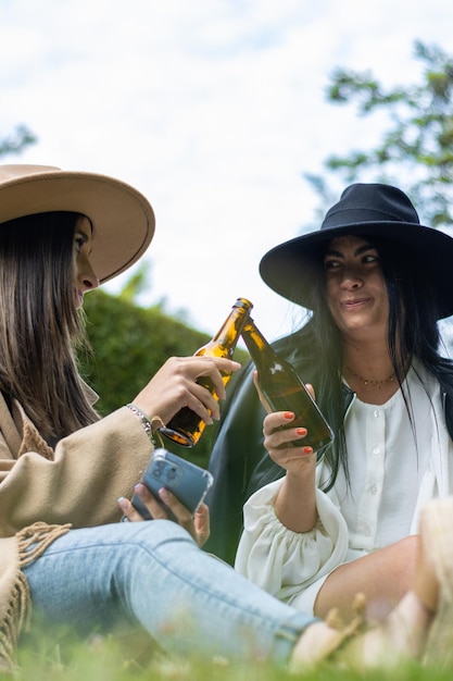 Portrait of two smiling young women in hats toasting with bottles of beer in the park