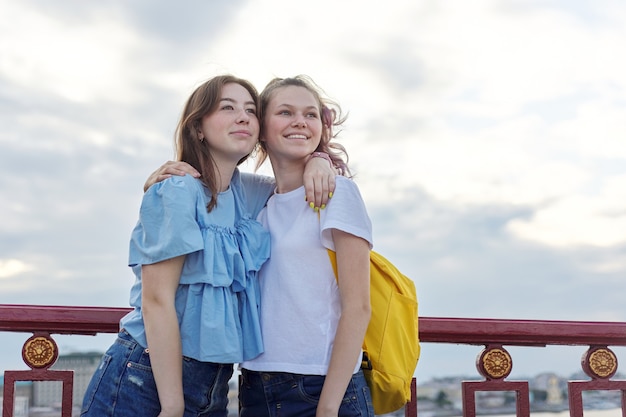 Portrait of two teenage girls standing on bridge over river, friends walk on summer sunny day. Friendship, lifestyle, youth, teens