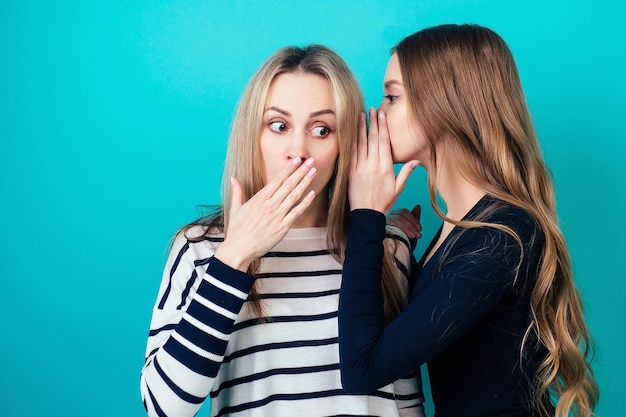 Photo portrait of two young attractive happy women with blonde hair and makeup eavesdrops whispers a secret (mystery) in the studio on a blue background. the concept of gossip and confidentiality