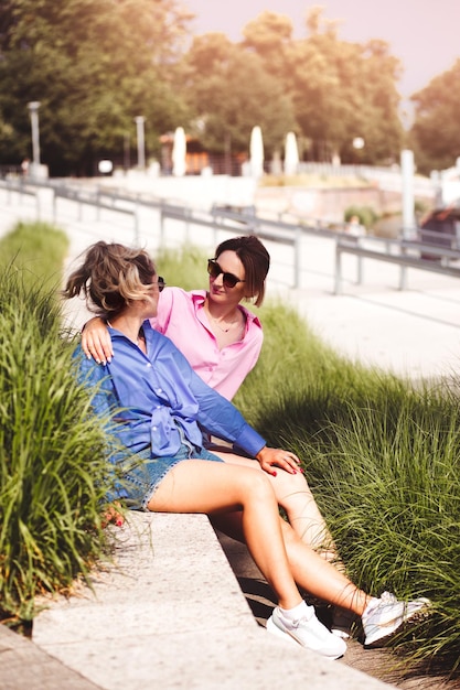 Portrait of two young beautiful smiling hipster female in trendy summer color clothes carefree women posing on river background Positive models having fun hugging