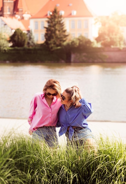 Portrait of two young beautiful smiling hipster female in trendy summer color clothes carefree women posing on river background Positive models having fun hugging