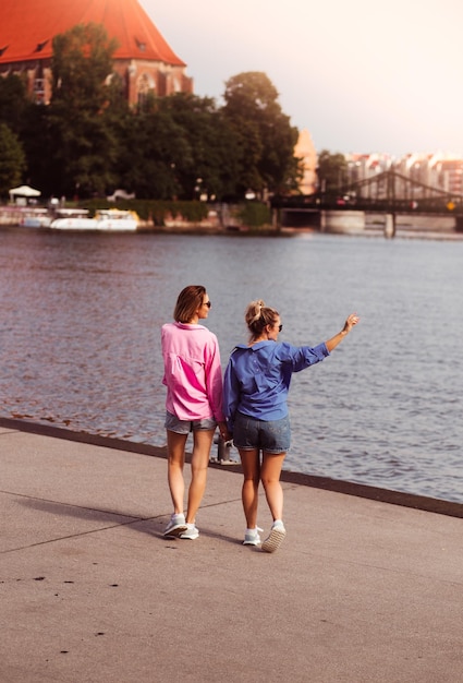 Portrait of two young beautiful smiling hipster female in trendy summer color clothes carefree women posing on river background Positive models having fun hugging