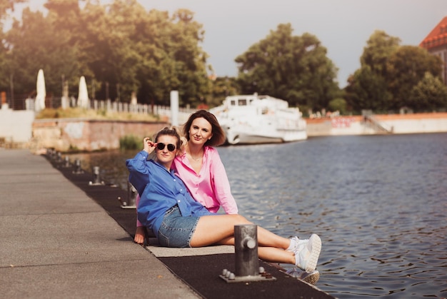 Portrait of two young beautiful smiling hipster female in trendy summer color clothes carefree women posing on river background Positive models having fun hugging