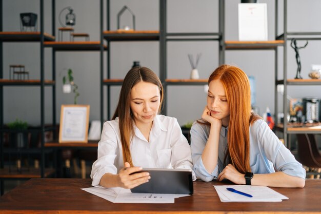 Portrait of two young business women working using digital tablet having meeting sitting at desk with job documents at office. Business partners using touchscreen computer for project discussion.