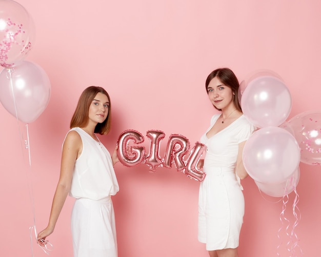 Portrait of two young cheerful friends dressed in a white holdings a balloons over pink background