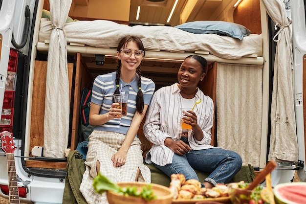 Portrait of two young girls drinking cocktails and smiling at camera while sitting in motorhome duri