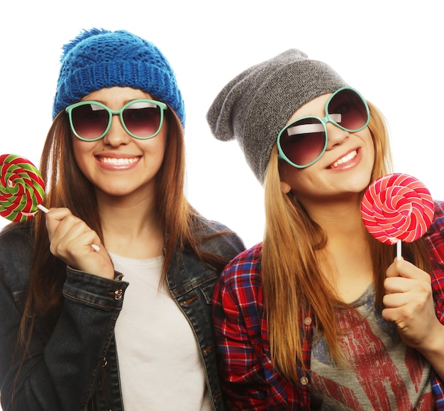 Portrait of two young pretty hipster girls wearing  hats and sunglasses holding candys. Studio portrait of two cheerful best friends having fun and making funny faces.