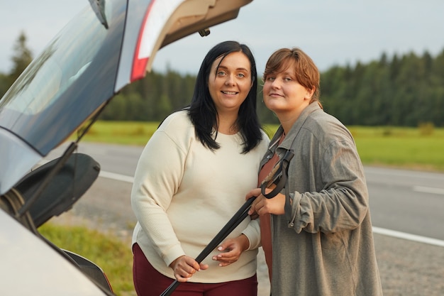 Portrait of two young women standing on the road and smiling at camera while travelling by car