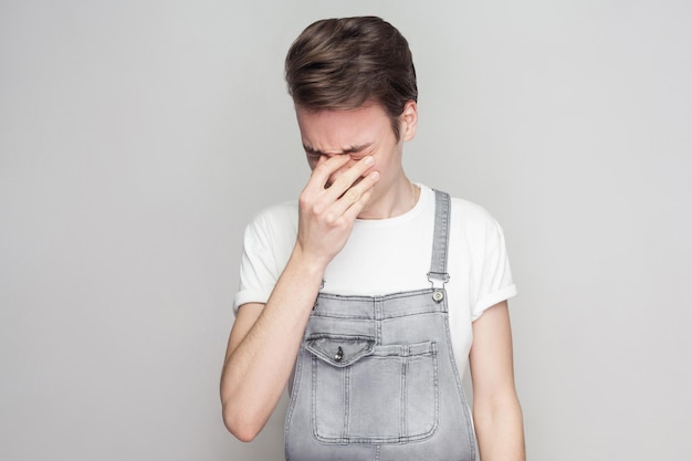 Portrait of unhappy broken heart young brunette man in casual style with white t-shirt and denim overalls standing, holding head down and crying. indoor studio shot, isolated on gray background.