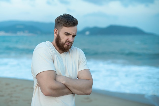 Portrait of unhappy depressed frozen trembling guy young shivering from cold sad upset man on the sea beach suffering from bad weather on summer vacation