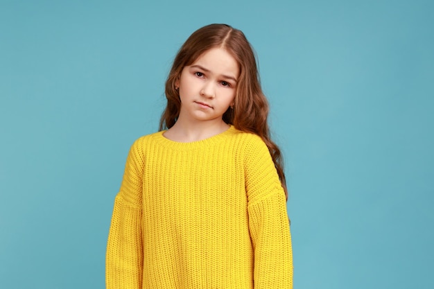 Portrait of unhappy little girl looking at camera with sad upset expression, being in bad mood, wearing yellow casual style sweater. Indoor studio shot isolated on blue background.