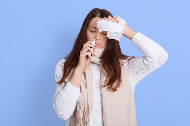 Portrait of unhappy young woman holding napkin and using nasal spray, touching her head, suffering from headache and fever, keeps eyes closed, wearing white sweater and scarf.