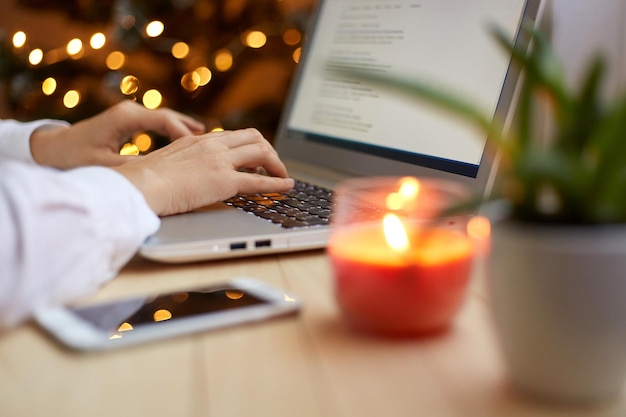 Portrait of unknown faceless woman typing on laptop keyboard while working in office interior decorated for Christmas holidays posing near xmas tree