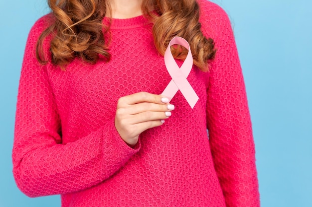 Portrait of unknown woman with wavy hair wearing pink pullover standing holding pink ribbon in front of chest, breast cancer, women health concept. Indoor studio shot isolated on blue background.