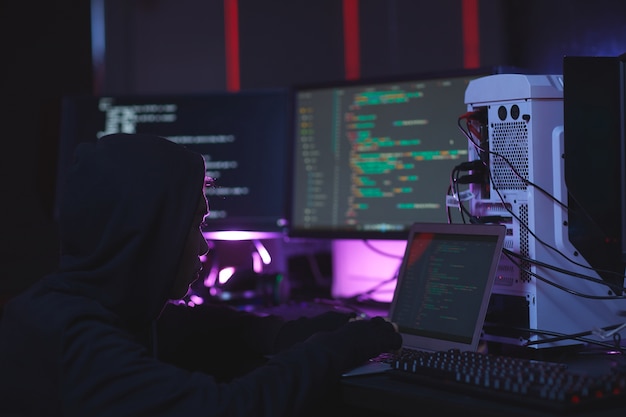 Portrait of unrecognizable hacker using computer equipment with programming code on screens in dark room, cyber security concept, copy space